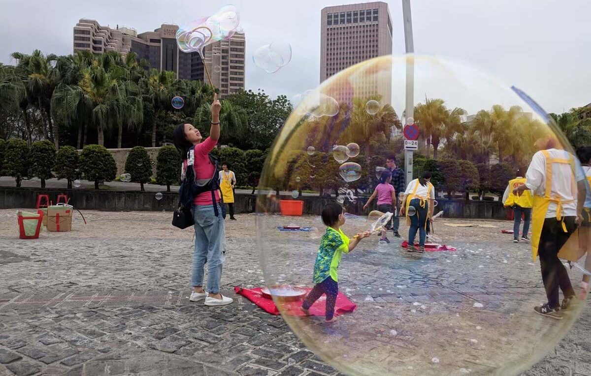 A photograph of a woman in Taiwan making soap bubbles in a public square with children playing along. A soap bubble is in the foreground, with the action in the background.