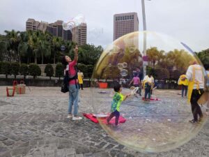A photograph of a woman in Taiwan making soap bubbles in a public square with children playing along. A soap bubble is in the foreground, with the action in the background.