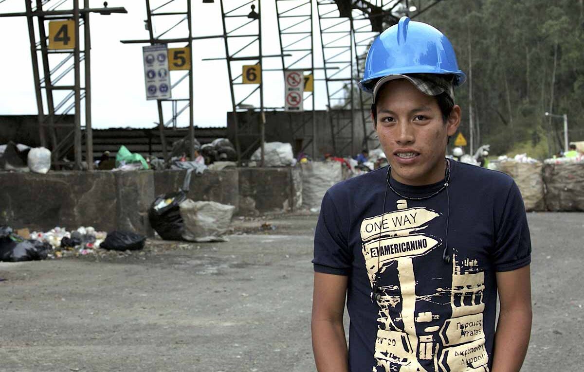 An image of a teenager, Marco, on the foreground, wearing a dark blue T-shirt, a cap and a helmet. In the background there is rubbish arranged in rows.