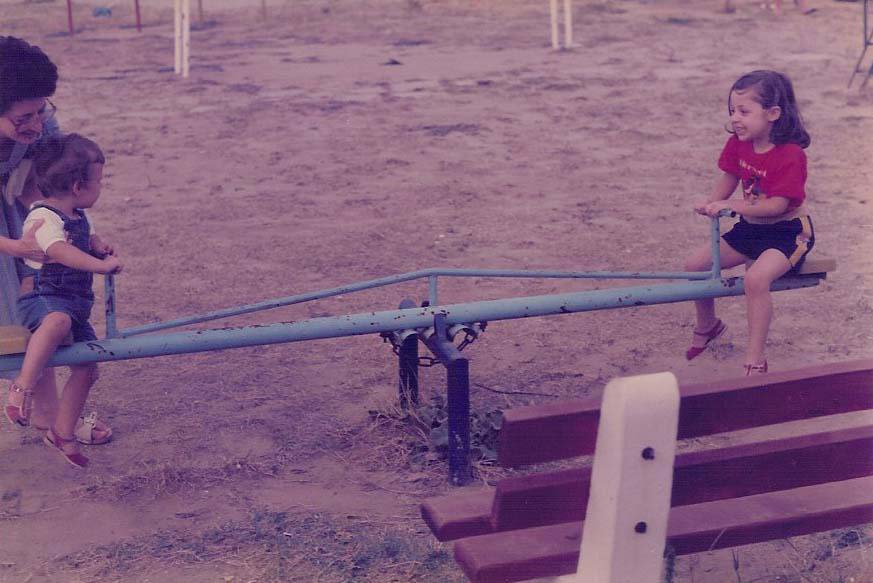 This photograph shows a smiley girl to the right of a seesaw, with a boy being lifted on the left side of the seesaw by their mother