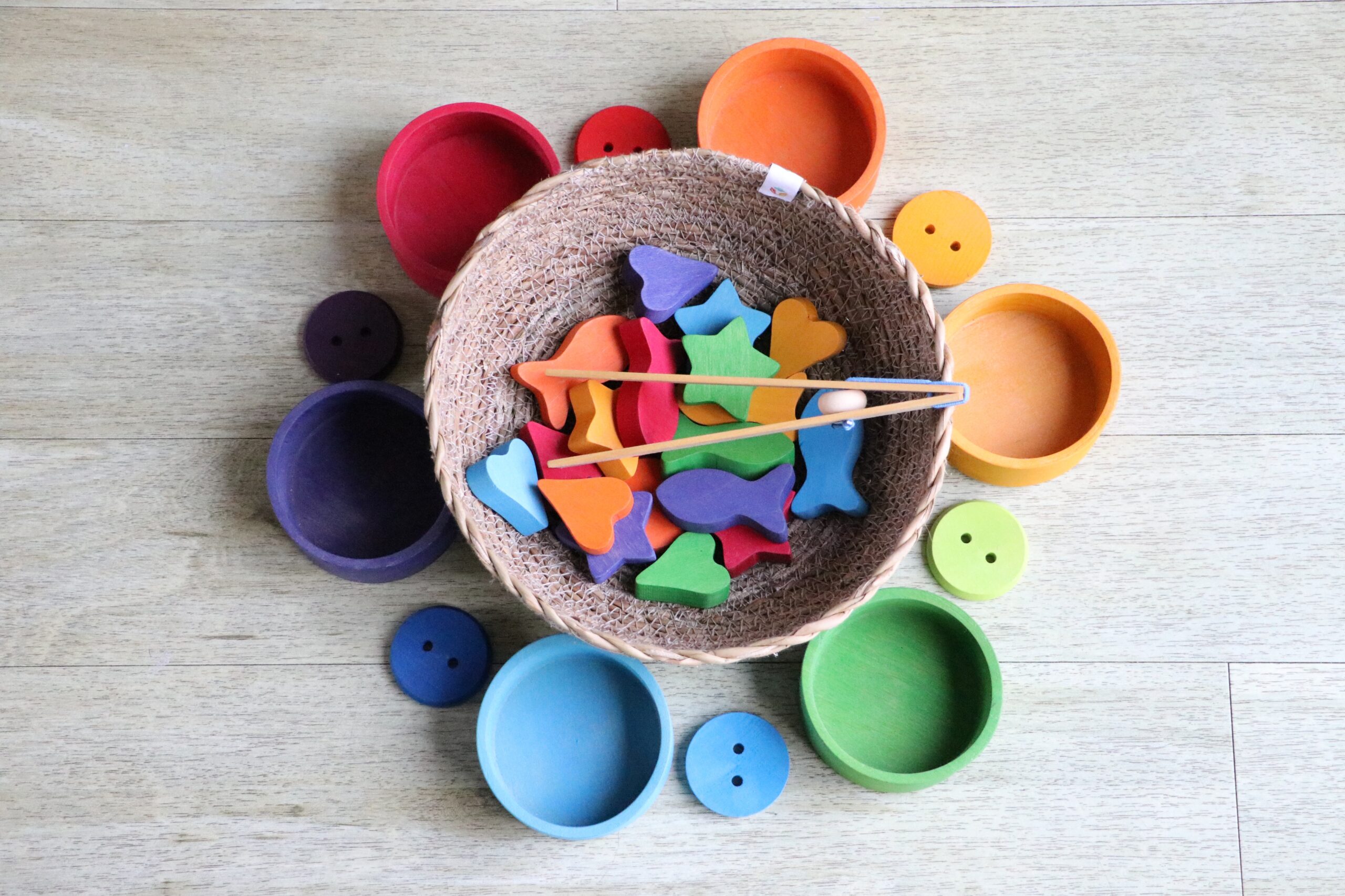 Round assorted-colors plastic cases. In the center one there are several wooden colorful figurines, shaped like fish, hearts and stars.