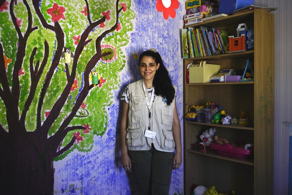 A smiling young woman standing against a wall with the drawing of a tree and a shelf with books and toys.